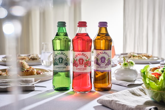 Drei Glasflaschen mit unterschiedlichen Limonaden auf einem gedeckten Tisch.