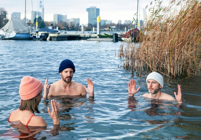 Drei Menschen in Badekleidung und mit Haube baden im kalten See