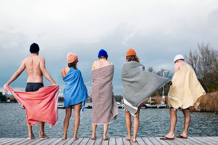 Fünf Menschen in Badekleidung und mit Haube stehen auf einem Steg vor einem Wasser in Badetücher gehüllt bevor sie eisbaden.