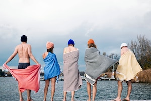 Fünf Menschen in Badekleidung und mit Haube stehen auf einem Steg vor einem Wasser in Badetücher gehüllt bevor sie eisbaden.