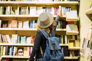 Bücherregal voll mit Büchern, Frau greift ein Buch aus dem Regal, junge Frau mit Jeansrucksack, lockigem Haar und Strohhut