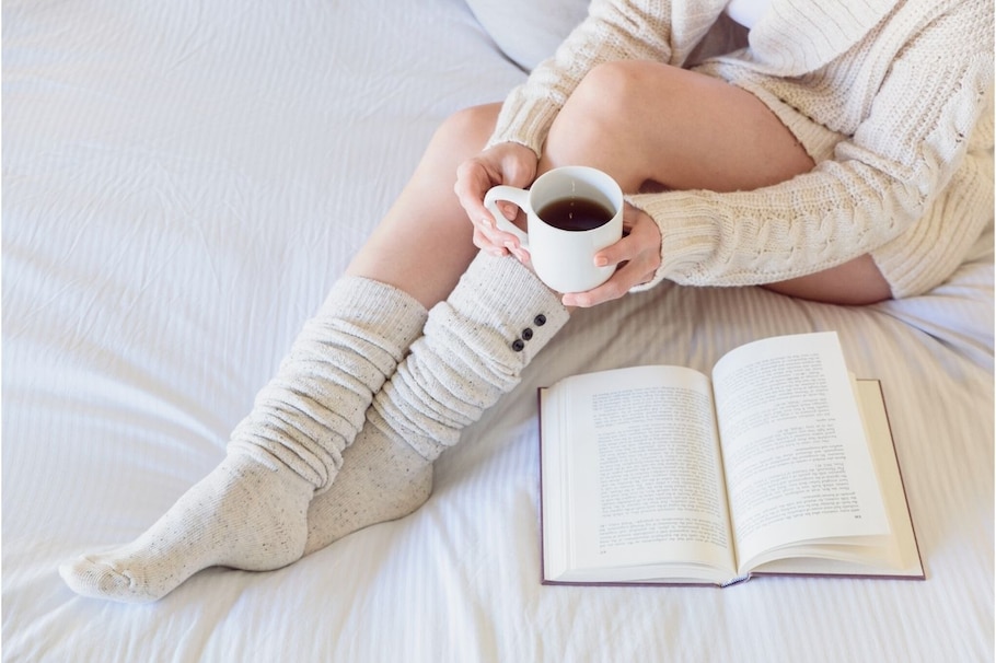Frauenbeine in Socken mit Essigpatscher, dazu Tee und Buch auf dem Bett
