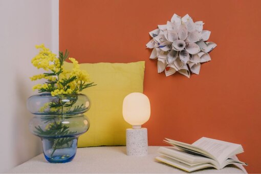 Papierblume auf oranger Wand, gelber Polster, Vase mit Blumen, aufgeschlagenes Buch und Lampe.