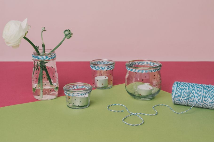 Vier Deko-Gläser mit Blume und Teelichtern. Im Hintergrund liegt eine Rolle Schnur.