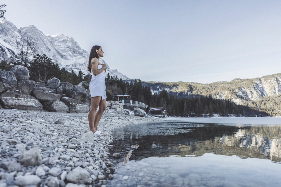 Frau in Badetuch gewickelt auf einem Steinstrand vor einem See. Im Hintergrund verschneite Berge.