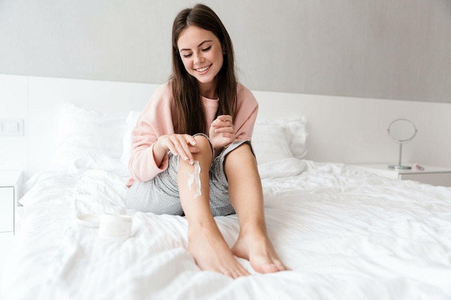 Frau im Pyjama auf dem Bett cremt sich Reibeisenhaut am Bein ein