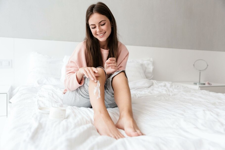 Frau im Pyjama auf dem Bett cremt sich Reibeisenhaut am Bein ein