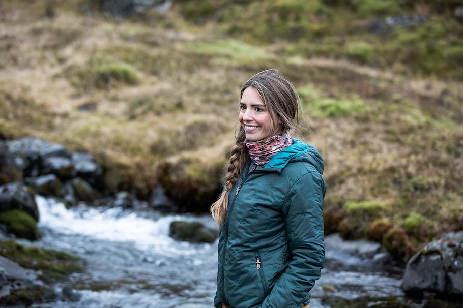 Eine Frau steht am Fluss und trägt ein Buff Tuch.