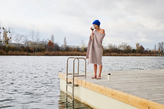 Eisbaden: Frau mit Mütze und Handtuch steht im Badeanzug am Wasser und trinkt Tee