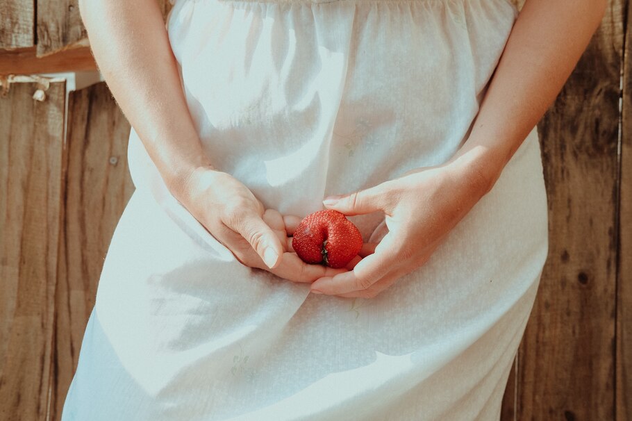 Detailaufnahme von einer Frau im weißen Kleid, die eine Erdbeere vor ihrem Schambereich in den Händen hält