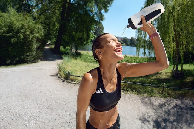 Ein Läuferin in einem städtischen Park an einem sonnigen Tag erfrischt ihr Gesicht mit Wasser aus ihrer Trinkflasche und genießt sichtlich die Abkühlung.